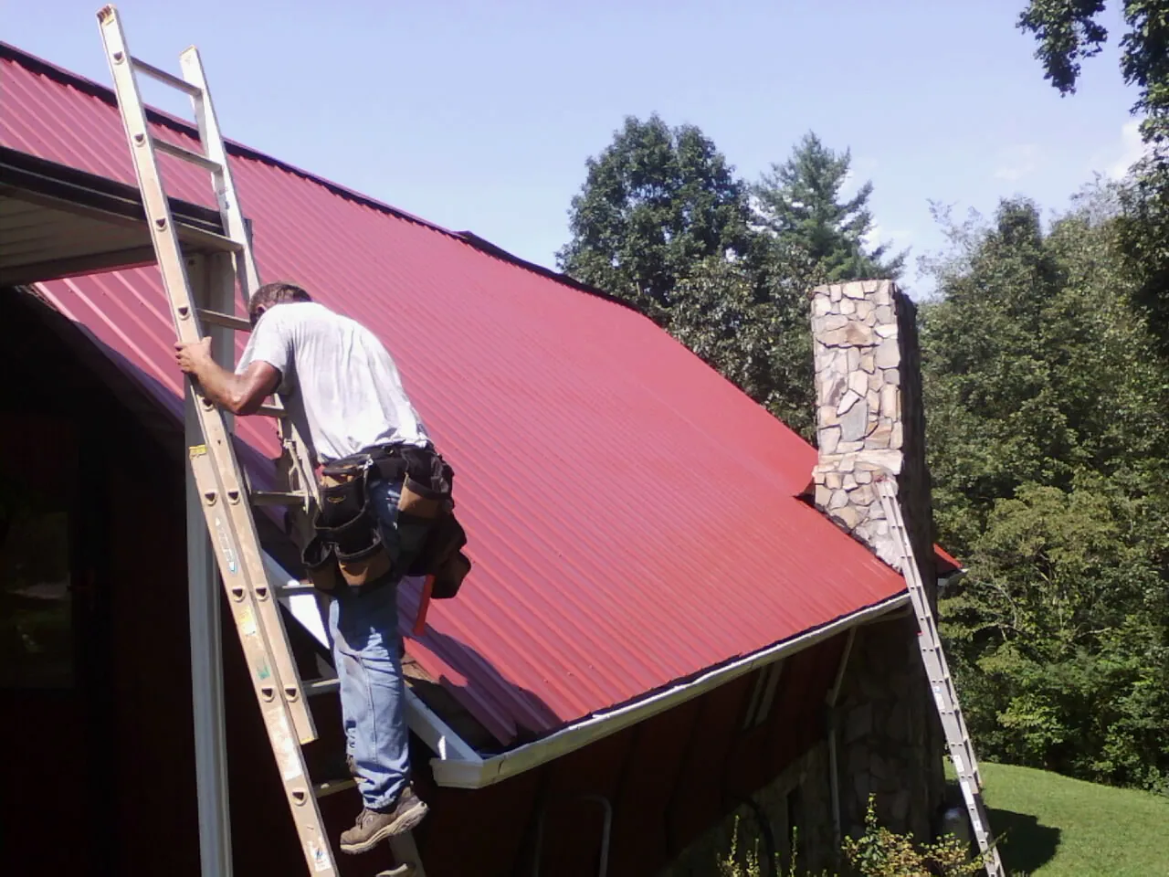 Roofer on a ladder beside a red metal roof and stone chimney — Soffit & Fascia Repair by Roofing Brothers in Upper Black Eddy